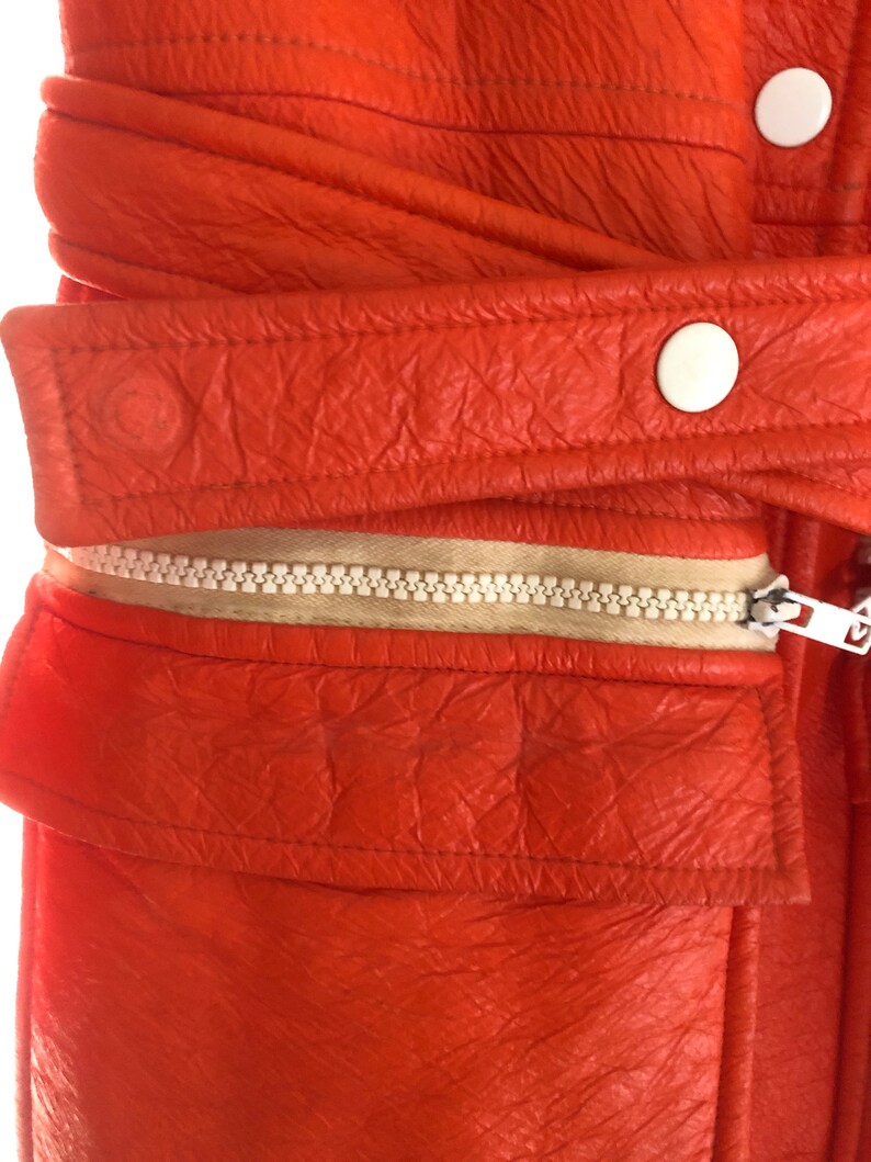 K&ouml;nnte beinhalten: Nahaufnahme einer leuchtend orangefarbenen Lederjacke mit Rei&szlig;verschluss und wei&szlig;en Kn&ouml;pfen. Die Jacke hat eine strukturierte Oberfl&auml;che und einen wei&szlig;en Rei&szlig;verschluss.