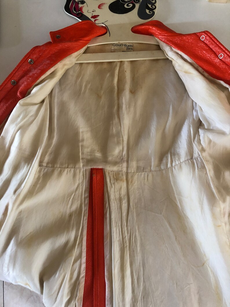 K&ouml;nnte beinhalten: Eine leuchtend orangefarbene Lederjacke mit wei&szlig;em Futter. Die Jacke ist auf links gedreht und zeigt das Futter und den Rei&szlig;verschluss. Das Etikett auf der Jacke lautet "Courreges Paris".