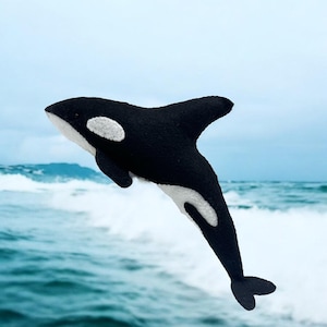 Puede incluir: Un peluche hecho a mano de una orca en blanco y negro, con vientre y parche ocular blancos. El juguete está situado sobre un fondo de olas del océano y un cielo nublado, creando una escena lúdica.