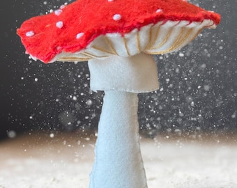 Peut inclure: Un ornement champignon en feutrine fait main avec un chapeau rouge vif à pois blancs brodés et un pied blanc. Le dessous du chapeau présente de fines coutures dorées, et une boucle en ficelle de jute est fixée au sommet pour suspendre.