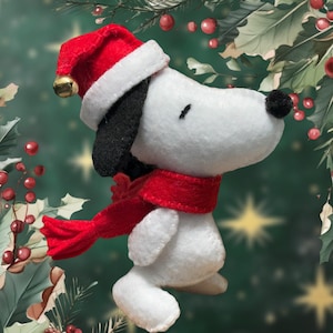 Puede incluir: Un perro de peluche blanco de fieltro que lleva un gorro de Papá Noel rojo con una campana y una bufanda roja. El perro está de pie sobre un fondo verde con bayas rojas.