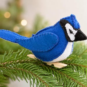 Peut inclure: Un ornement en feutrine représentant un geai bleu, composé de pièces de tissu bleu, blanc et noir assemblées par des coutures visibles. L'oiseau présente un bec noir pointu, une poitrine blanche et une crête bleue sur un socle à pince en bois.