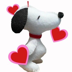 Puede incluir: Una figura de Snoopy de fieltro blanco con una oreja negra y un collar rojo, sosteniendo tres corazones rojos con contornos rosas. La figura está sobre un fondo blanco. La imagen tiene un tema de San Valentín.