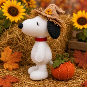 May include: A white felt Snoopy figurine with a black ear, a red collar, and a tan hat adorned with a small yellow flower. Beside the figurine sits a small orange felt pumpkin with green leaves and curly vines, all crafted from felt material.