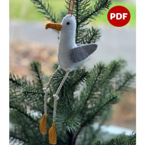 Puede incluir: Un adorno de gaviota de fieltro con un ala gris, un pico naranja y un cuerpo blanco. El adorno está colgado de una cuerda roja en una rama de pino verde. La palabra "PDF" está escrita en letras blancas sobre un círculo rojo.
