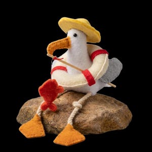Könnte beinhalten: Eine Möwenfigur aus Filz mit gelbem Hut und weißem Rettungsring mit roten Streifen. Die Möwe hat einen orangefarbenen Schnabel, graue Flügel und orangefarbene Schwimmfüße und hält eine hölzerne Angelrute mit einem kleinen roten Filzfisch.