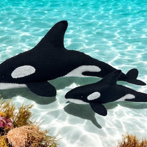 May include: Two plush killer whale toys, one large and one small, are displayed in a shallow, clear blue water setting. The whales are black with white markings and fins. The scene includes coral and seaweed.