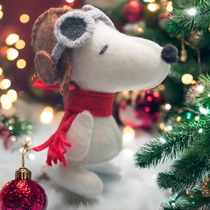 Puede incluir: Un perro de peluche blanco de fieltro que lleva un gorro de aviador marrón con gafas y una bufanda roja. El perro está de pie frente a un árbol de Navidad con luces y adornos.