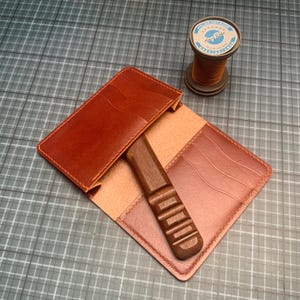 May include: A handmade leather wallet in shades of brown, with a wooden tool and a spool of thread. The wallet is open, revealing interior pockets and a light tan lining. The wooden tool has a carved handle. The spool of thread is labeled with text.