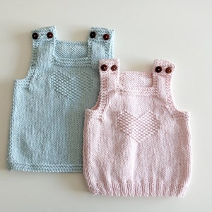 May include: Two knitted baby vests with heart designs. One vest is light blue and the other is pink.
