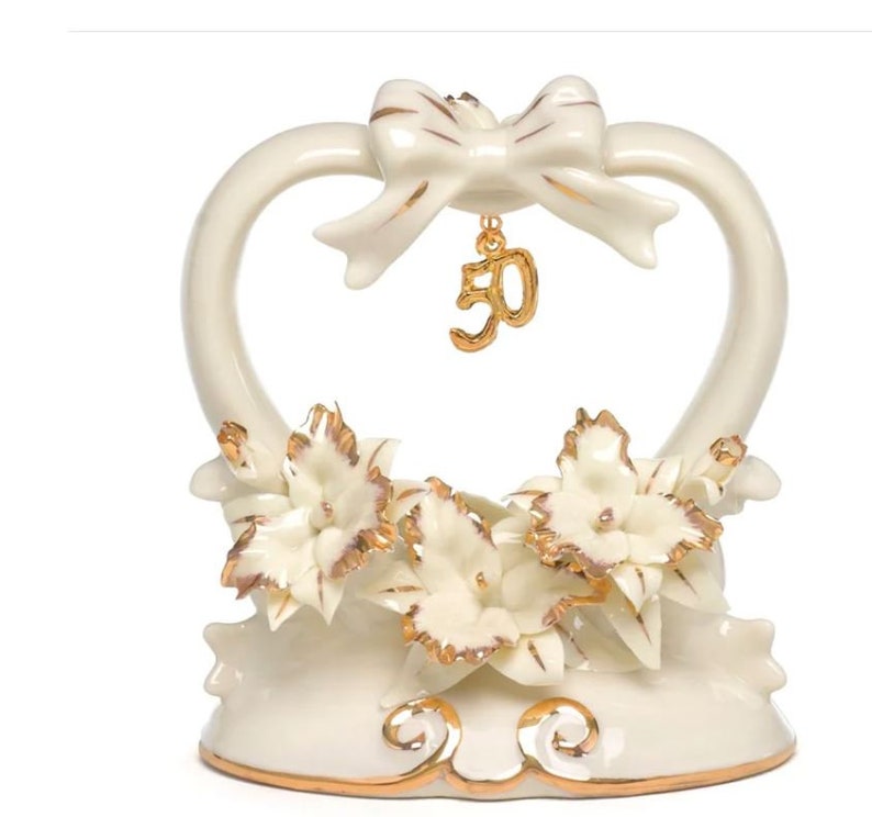 May include: Ivory-colored ceramic cake topper with a heart-shaped design. Features a bow at the top and a dangling gold number 50 charm. The base is adorned with white flowers with gold accents, perfect for a 50th anniversary celebration.