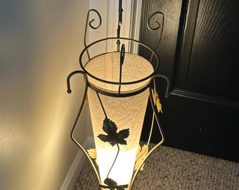 May include: A vintage floor lamp with a cream-colored shade and a metal frame. The lamp features decorative leaf accents and a three-legged base. The shade is illuminated, casting a warm glow.