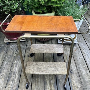 May include: A rolling cart featuring a rectangular wooden top surface with a warm brown finish. The frame is constructed from gold-toned metal tubing with two lower shelves that have a textured, metallic finish. The cart includes four small wheels.
