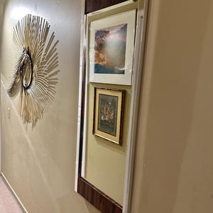 May include: A rectangular mirror with a dark wood frame and a silver trim. The mirror is hanging on a wall and has two framed pictures inside. The top picture is a landscape scene with a mountain range and a cloudy sky. The bottom picture is a framed print of a scene with people and trees.