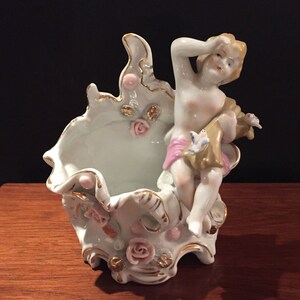 May include: A white porcelain figurine of a cherub sitting in a decorative planter. The planter is adorned with gold accents and pink flowers.