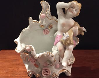 May include: A white porcelain figurine of a cherub sitting in a decorative planter. The planter is adorned with gold accents and pink flowers.