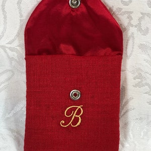 May include: A red fabric pouch with a snap closure. The pouch has a flap and is lined with a shiny red material. A gold embroidered letter "B" is on the front.