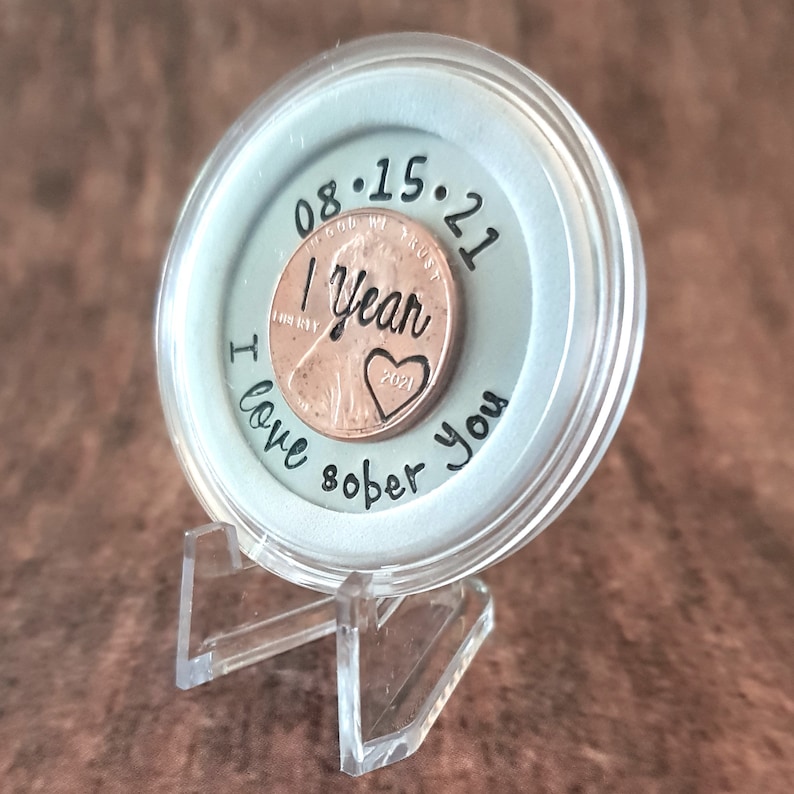 May include: A clear plastic coin holder with a silver background and a copper penny inside. The penny has the date 08.15.21 and the words "1 year" and "I love sober you" engraved on it.