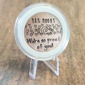 May include: A round, copper-colored coin with the text "365 Moons" and "We're so proud of you!" surrounded by stars and a crescent moon. The coin is displayed on a clear plastic stand.