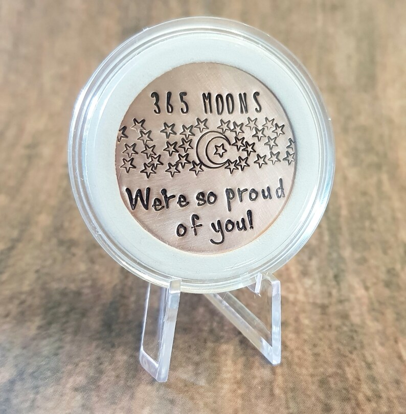May include: A round, copper-colored coin with the text "365 Moons" and "We're so proud of you!" surrounded by stars and a crescent moon. The coin is displayed on a clear plastic stand.