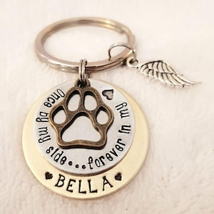 May include: Silver and gold tone keychain with a paw print charm, a small angel wing charm, and a round tag that reads "Once by my side...forever in my heart" with the name "BELLA" on the bottom.