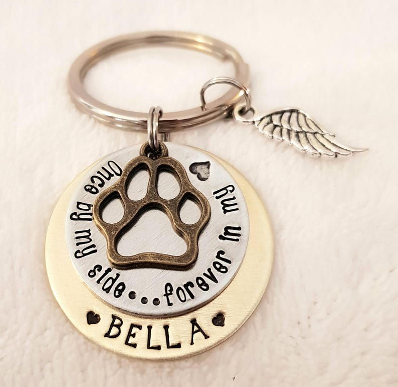 May include: Silver and gold tone keychain with a paw print charm, a small angel wing charm, and a round tag that reads "Once by my side...forever in my heart" with the name "BELLA" on the bottom.