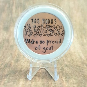 May include: A copper-colored coin with the text "365 Moons" and "We're so proud of you!" surrounded by stars and a crescent moon. The coin is displayed on a clear plastic stand.