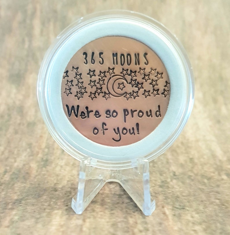 May include: A copper-colored coin with the text "365 Moons" and "We're so proud of you!" surrounded by stars and a crescent moon. The coin is displayed on a clear plastic stand.