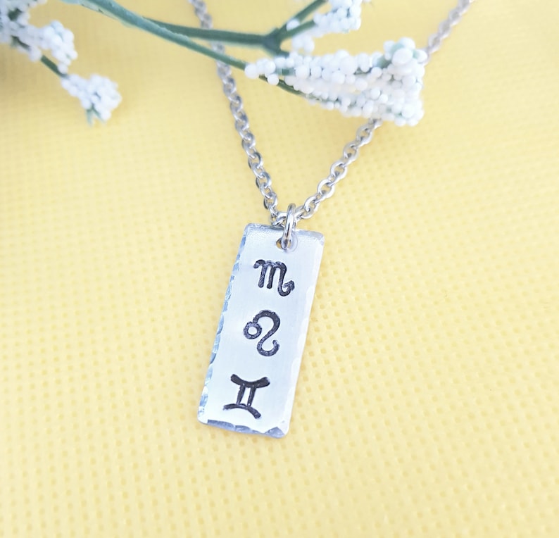 May include: Silver necklace with a rectangular tag pendant featuring the astrological symbols for Scorpio, Leo, and Gemini.