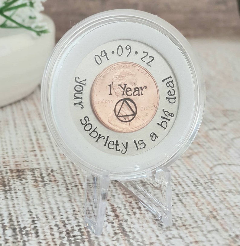 Op de afbeelding: Een heldere, ronde displaycase met een koperen penny. De penny is gestempeld met "1 Year" en een AA-symbool. De case is gegraveerd met "Your sobriety is a big deal" en de datum "04.09.22". De case staat op een heldere standaard.