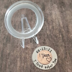 May include: A silver coin with the date 08.15.21 engraved around a copper penny with the words "1 Year Sober" and a heart. The coin also has the words "I love you" engraved around the penny.