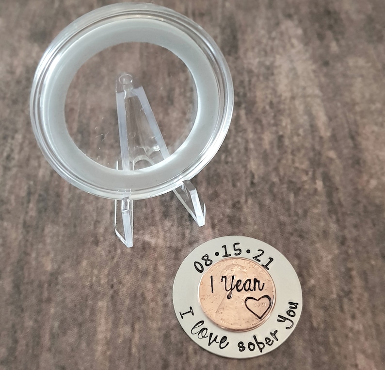 May include: A silver coin with the date 08.15.21 engraved around a copper penny with the words "1 Year Sober" and a heart. The coin also has the words "I love you" engraved around the penny.
