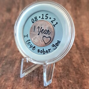 May include: A clear plastic coin holder with a copper penny inside. The penny has the date 08.15.21 and the words "1 Year Sober" engraved on it. The holder has the words "I love sober you" written on it.