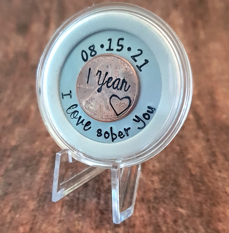 May include: A clear plastic coin holder with a copper penny inside. The penny has the date 08.15.21 and the words "1 Year Sober" engraved on it. The holder has the words "I love sober you" written on it.