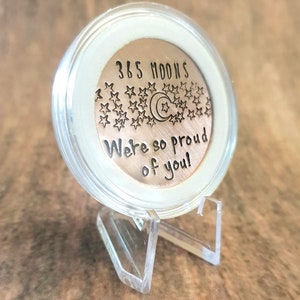 May include: A copper coin with the text "365 Moons We're so proud of you!" and a crescent moon and stars design. The coin is displayed in a clear plastic stand.