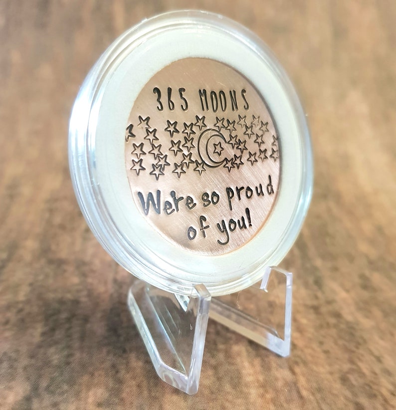 May include: A copper coin with the text "365 Moons We're so proud of you!" and a crescent moon and stars design. The coin is displayed in a clear plastic stand.