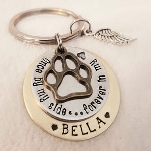 May include: A silver and gold keychain with a paw print charm and the text "Once by my side...forever in my heart" and the name "BELLA".