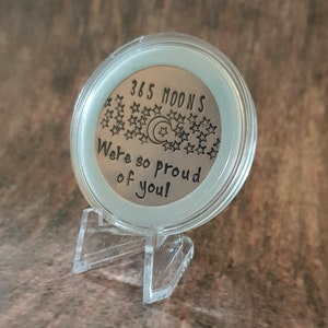 May include: A silver coin with the text "365 Moons We're so proud of you!" surrounded by stars and a crescent moon. The coin is on a clear plastic stand.