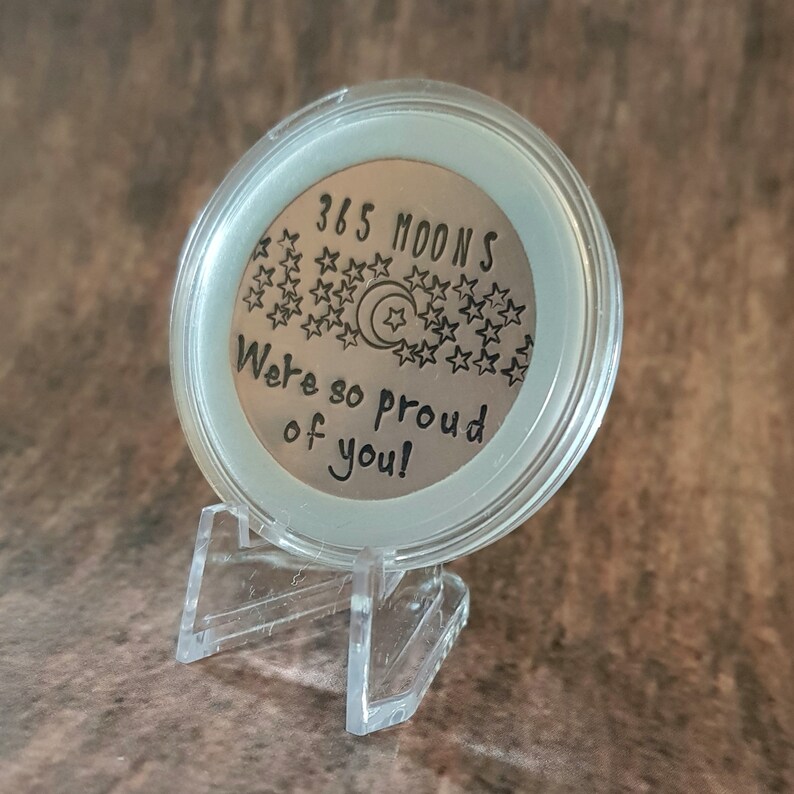 May include: A silver coin with the text "365 Moons We're so proud of you!" surrounded by stars and a crescent moon. The coin is on a clear plastic stand.