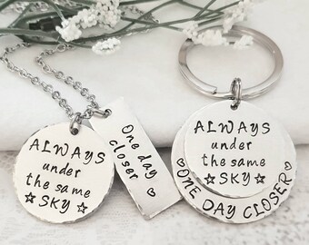 May include: Two silver tone keychains with a round charm and a rectangular charm. The round charm says "ALWAYS under the same ☆SKY☆" and the rectangular charm says "One day closer." The other keychain has a round charm that says "ALWAYS under the same ☆SKY☆" and a rectangular charm that says "One day closer."