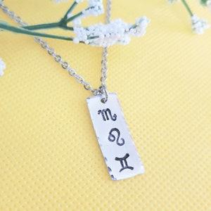 May include: A silver necklace with a rectangular pendant featuring the astrological symbols for Scorpio, Leo, and Gemini.