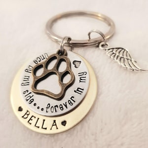 May include: Silver and gold tone keychain with a paw print charm, a small heart charm, and a small angel wing charm. The keychain has the text "Once you enter my life...forever in my heart" and "BELLA" on it.