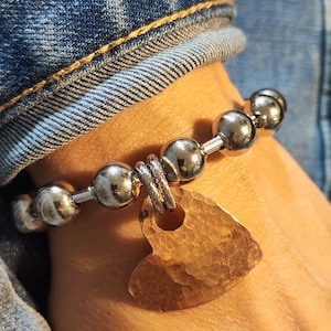May include: Silver beaded bracelet with a hammered copper heart charm. The bracelet features large, round silver beads connected by smaller silver bars. The heart charm has a textured surface and a warm copper color.