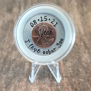 May include: A clear plastic coin holder with a copper penny inside. The penny has the date 08.15.21 and the words "1 year" and "2021" engraved on it. The holder has the words "I love sober you" written around the edge.