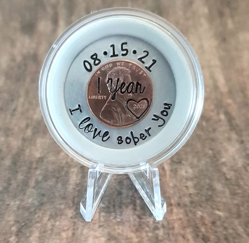 May include: A clear plastic coin holder with a copper penny inside. The penny has the date 08.15.21 and the words "1 year" and "2021" engraved on it. The holder has the words "I love sober you" written around the edge.