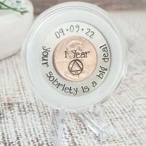 Op de afbeelding: Een heldere, ronde displaycase met een penny met de tekst "1 Year" en een AA-symbool. De case staat op een heldere standaard. De tekst "Your sobriety is a big deal" en de datum "04.09.22" zijn ook zichtbaar.