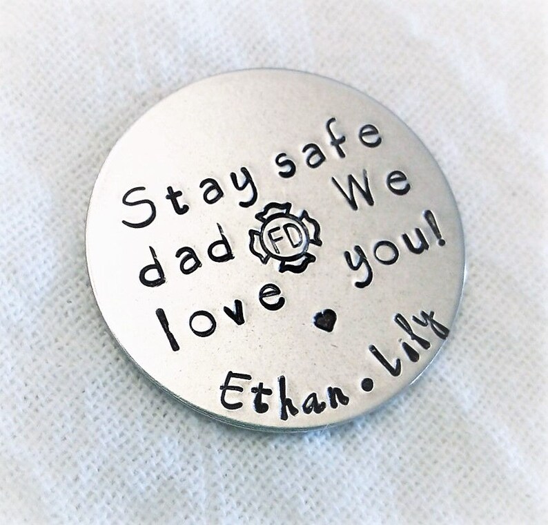 May include: Silver round disc with the text "Stay safe dad We love you!" and a firefighter's badge with the letters "FD" inside. The text "Ethan. Lily" is also on the disc.