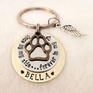 May include: A silver and gold keychain with a paw print charm, a small angel wing charm, and the text "Once by my side...forever in my heart Bella".