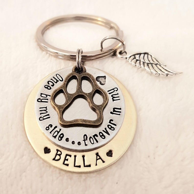 May include: A silver and gold keychain with a paw print charm, a small angel wing charm, and the text "Once by my side...forever in my heart Bella".