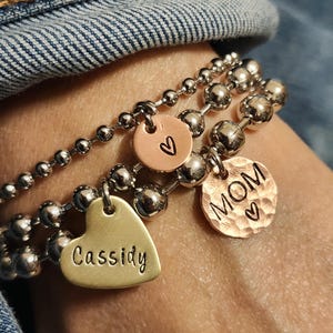 May include: Three silver beaded bracelets with personalized charms. One charm is a gold heart with the name "Cassidy", another is a copper circle with a heart, and the third is a copper circle with the word "MOM" and a heart.